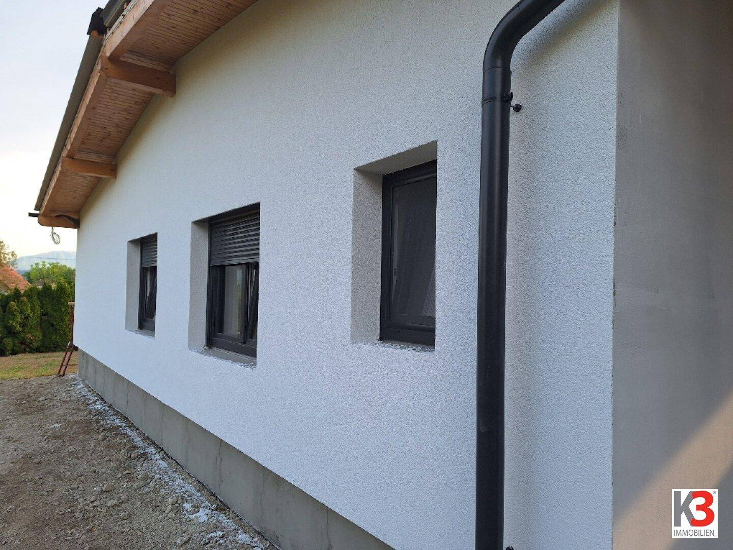 Seitenansicht eines Neubaus mit weißer Fassade, dunklen Fenstern und Fallrohr.