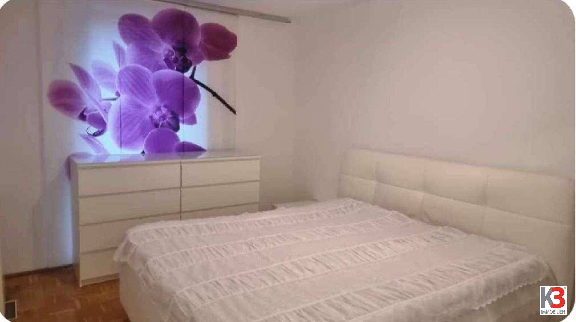 Schlafzimmer mit weißem Bett, Kommode und einem großen, farbenfrohen Orchideenbild als Wanddekoration.