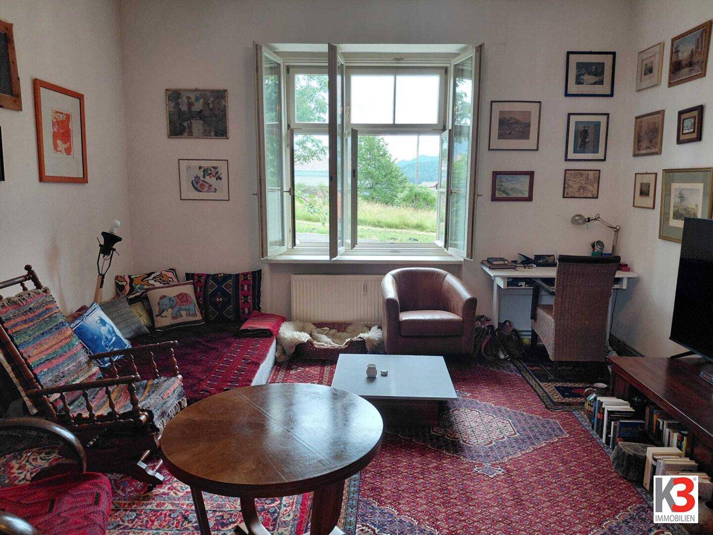 Gemütliches Wohnzimmer mit Teppichboden, vielen Bildern und einem weiten Blick ins Grüne.