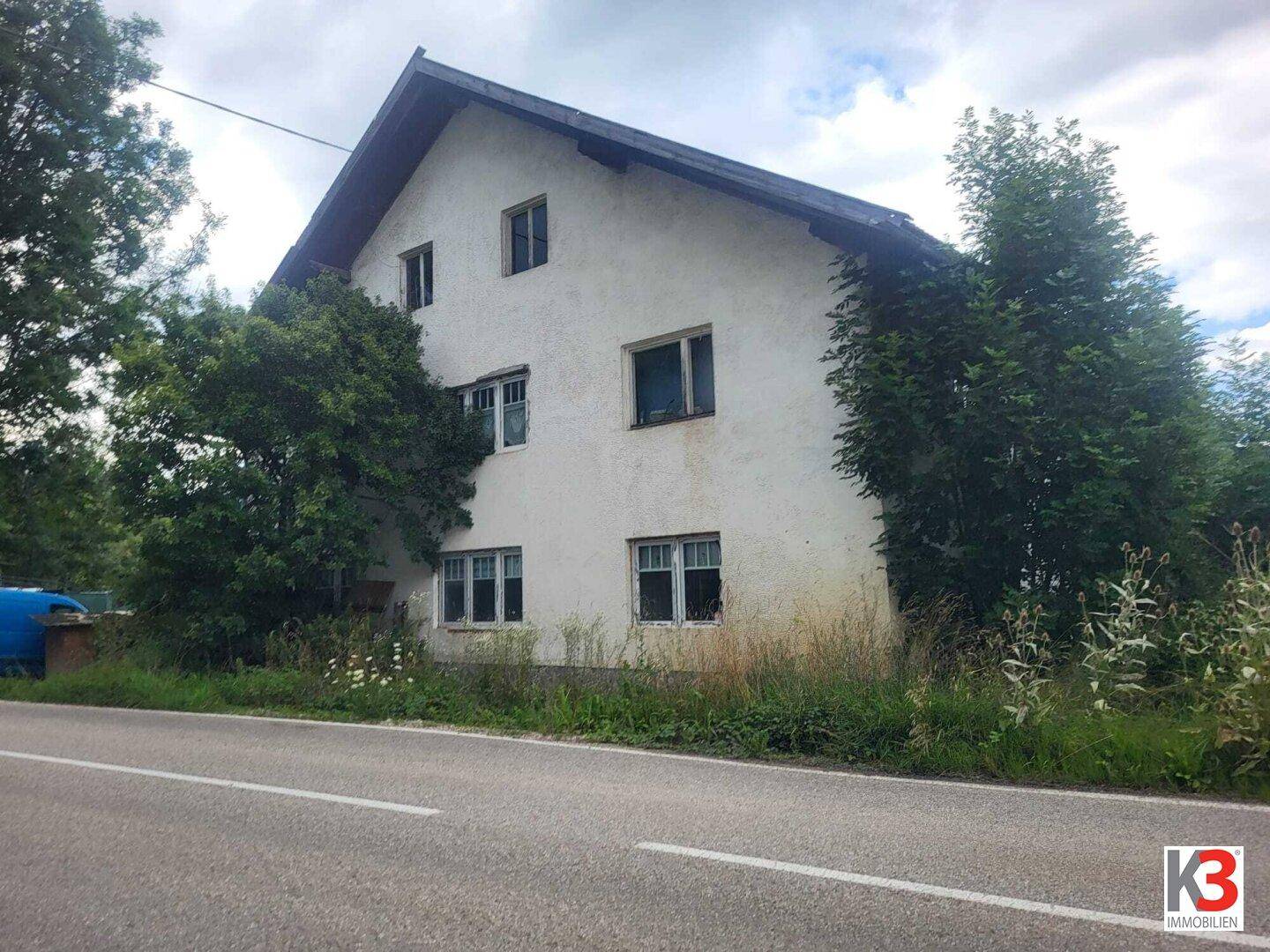 Frontansicht eines älteren Wohnhauses mit mehreren Fenstern und einem Satteldach, direkt an einer Landstraße gelegen.