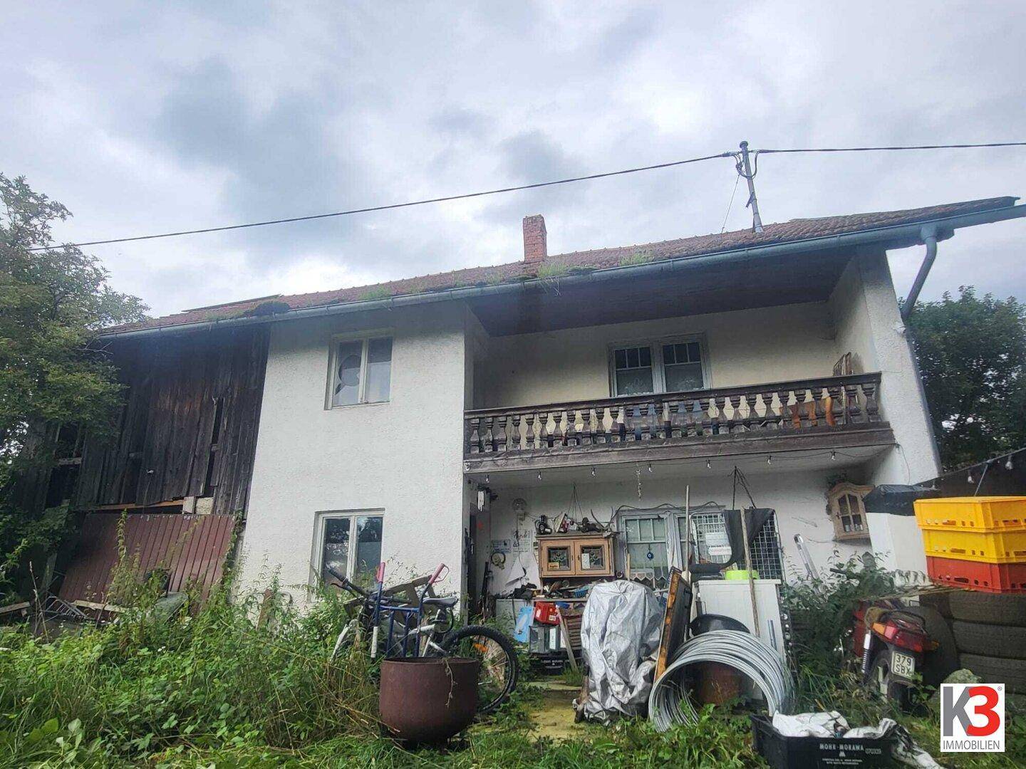 Rückseite eines älteren Hauses mit einem Holzbalcon und viel Unordnung im Gartenbereich.