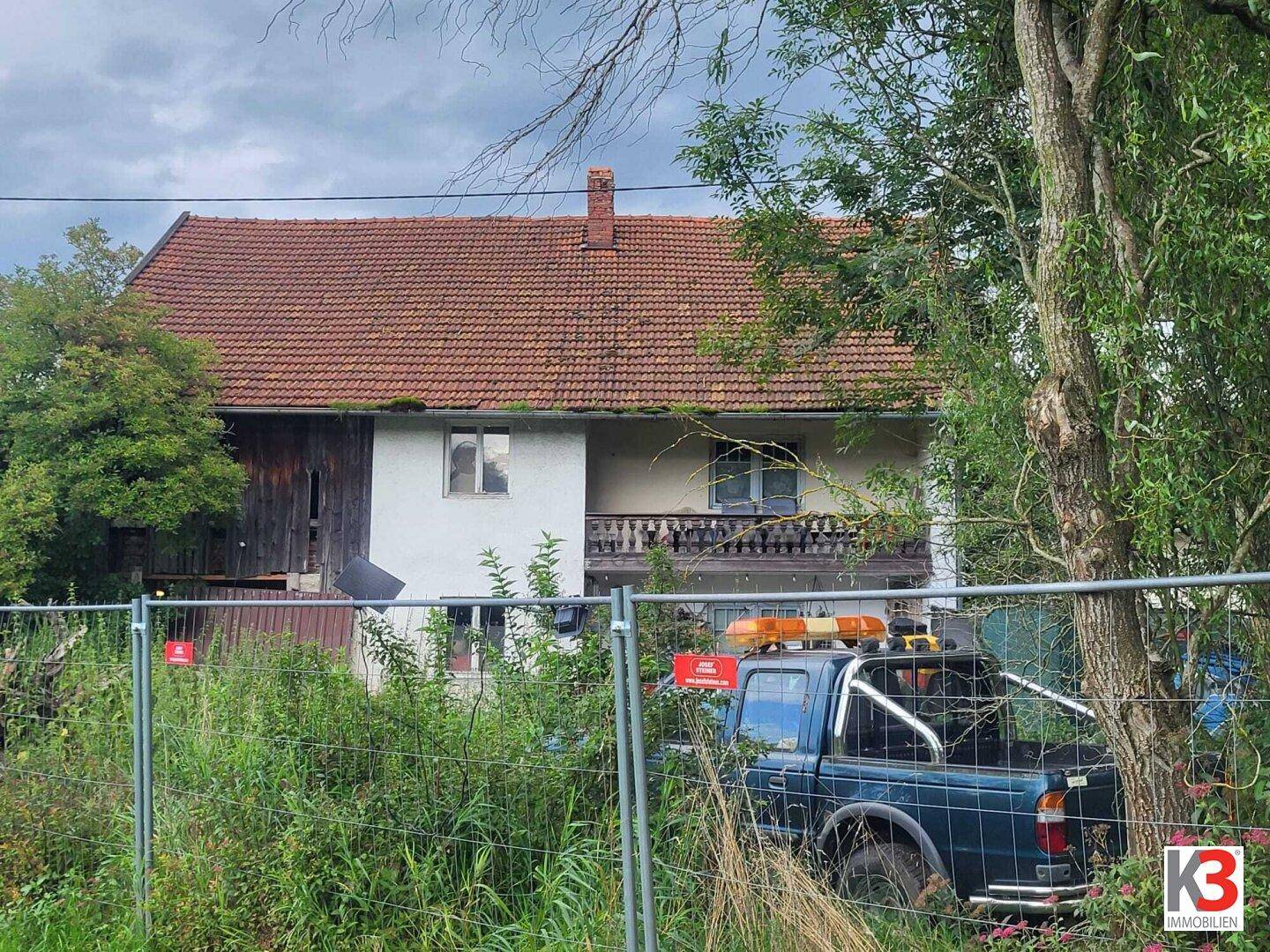 Älteres Wohnhaus mit Satteldach und Balkon, teilweise verdeckt durch einen Bauzaun und Vegetation.