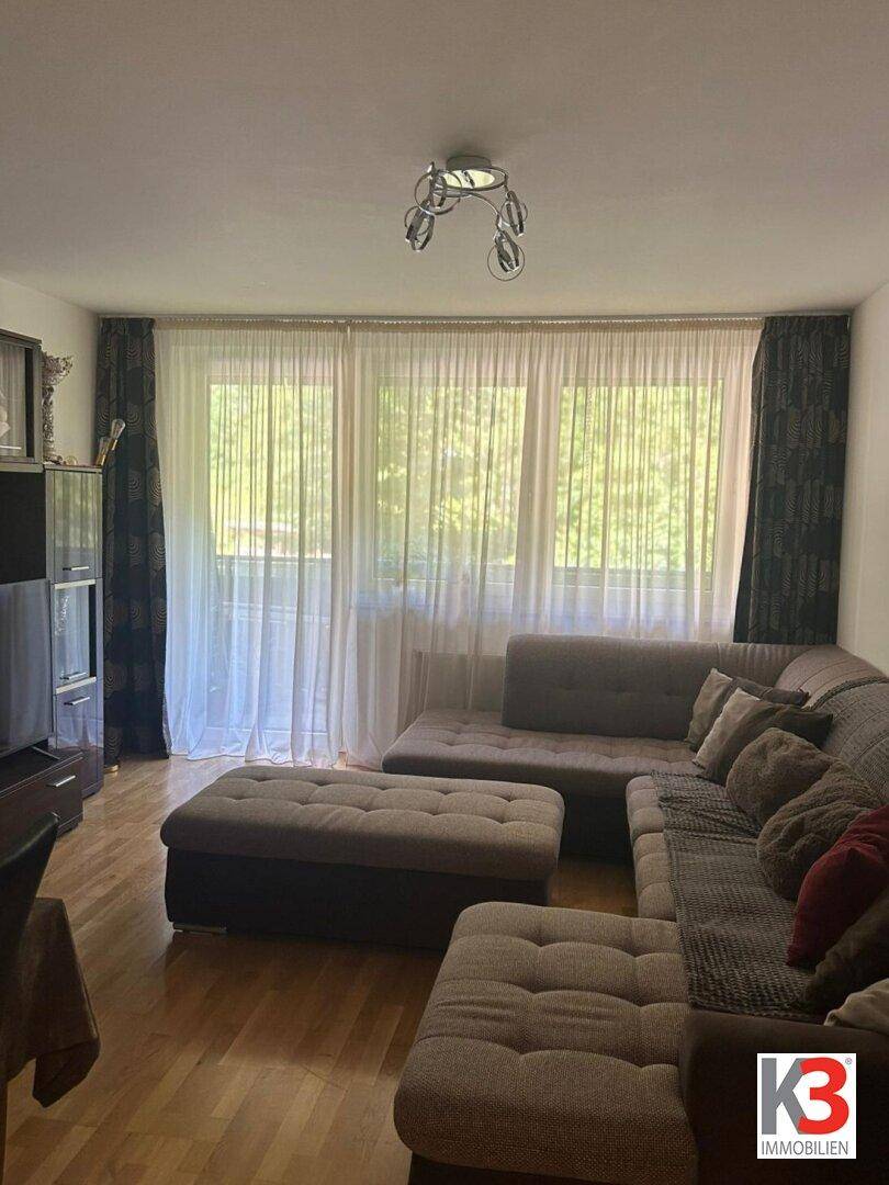 Helles Wohnzimmer mit großem Sofa, Holzboden und Zugang zu einem Balkon mit Grünblick.