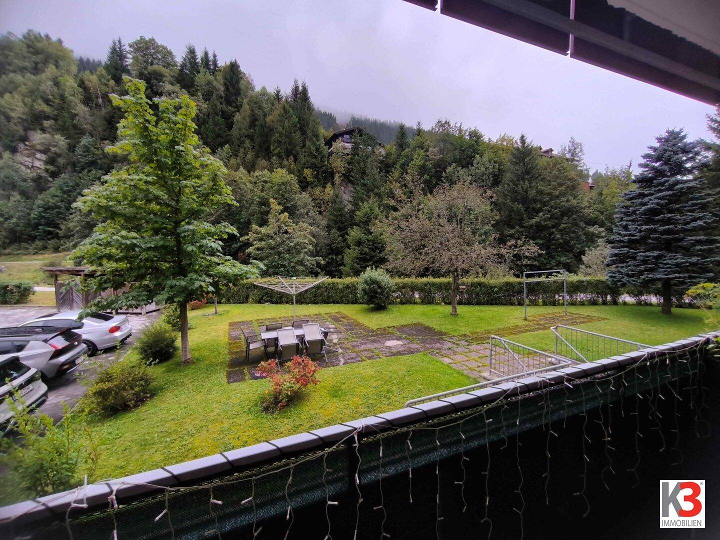 Balkon mit Blick auf den Gemeinschaftsgarten, umgeben von Bäumen und Bergen.