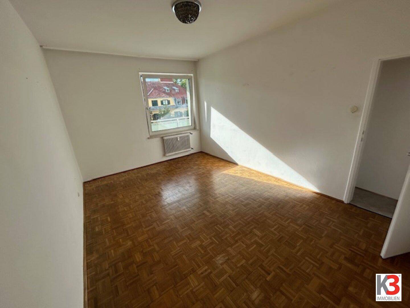 Heller, leerer Raum mit Fischgrätparkettboden und einem Fenster, das Tageslicht hereinlässt.