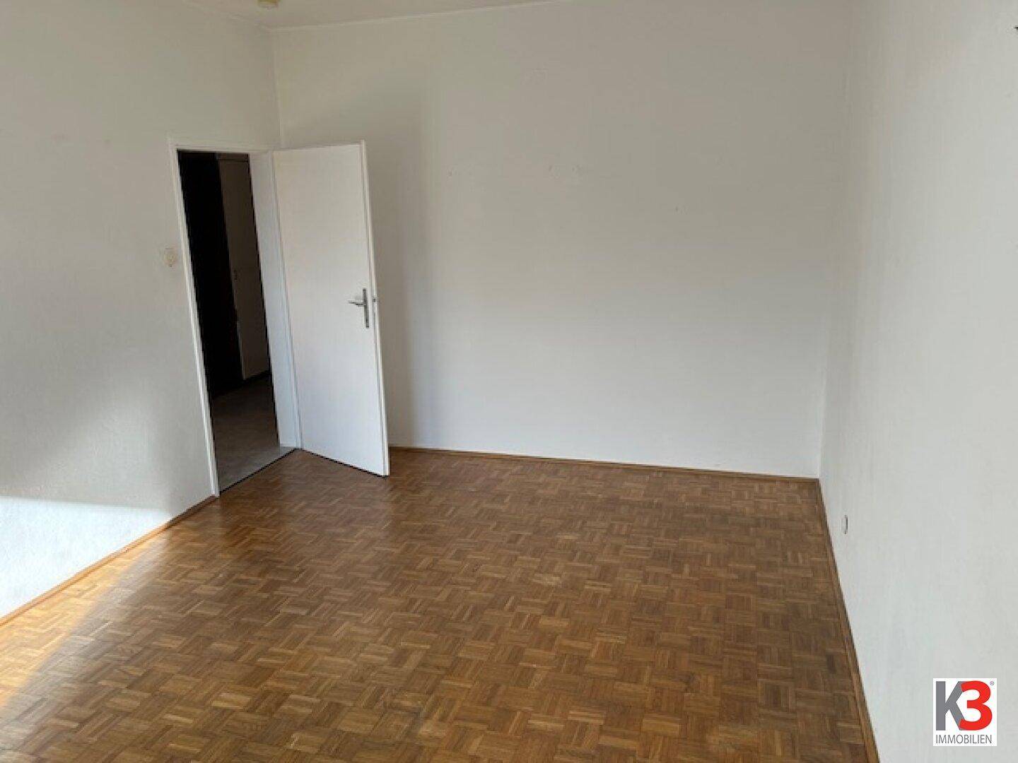 Leeres Zimmer mit Parkettboden und einer offenen Tür, die den Zugang zu einem weiteren Bereich zeigt.