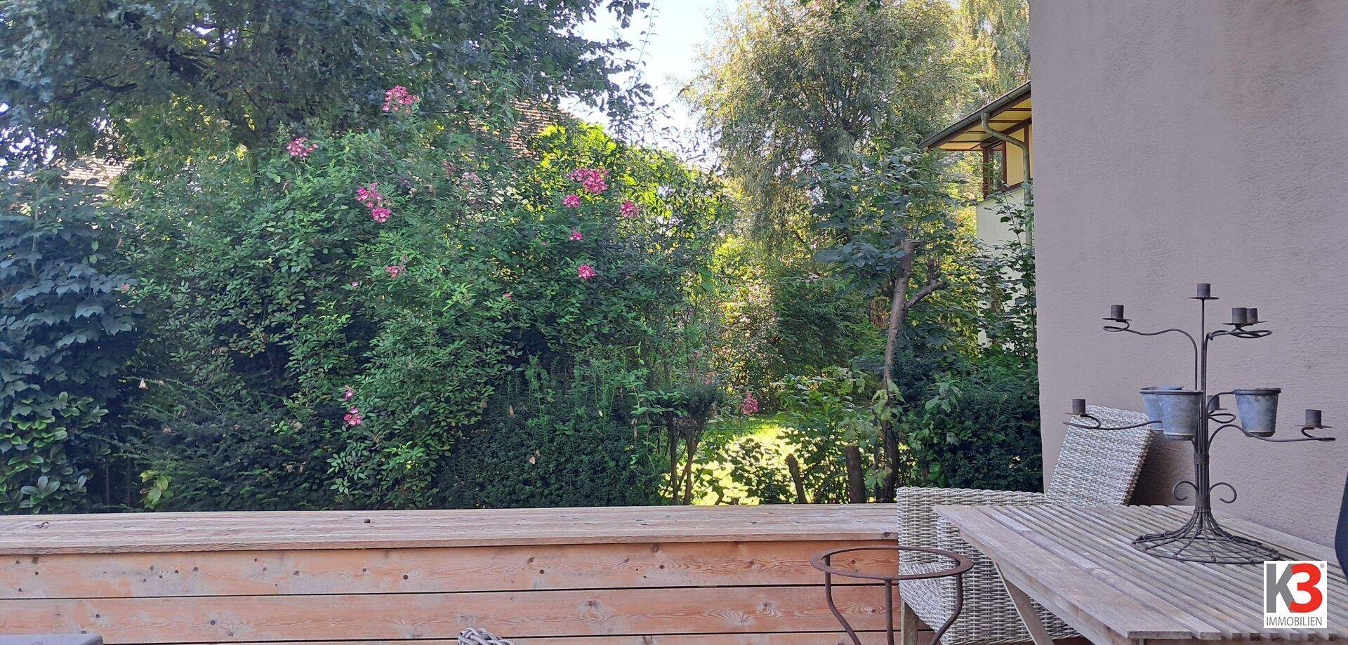 Blick von der Terrasse auf üppiges Grün, blühende Büsche und hohe Bäume.