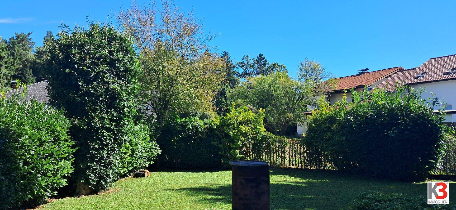 Gepflegter Garten mit großem Rasen, umgeben von Hecken und Bäumen unter blauem Himmel.