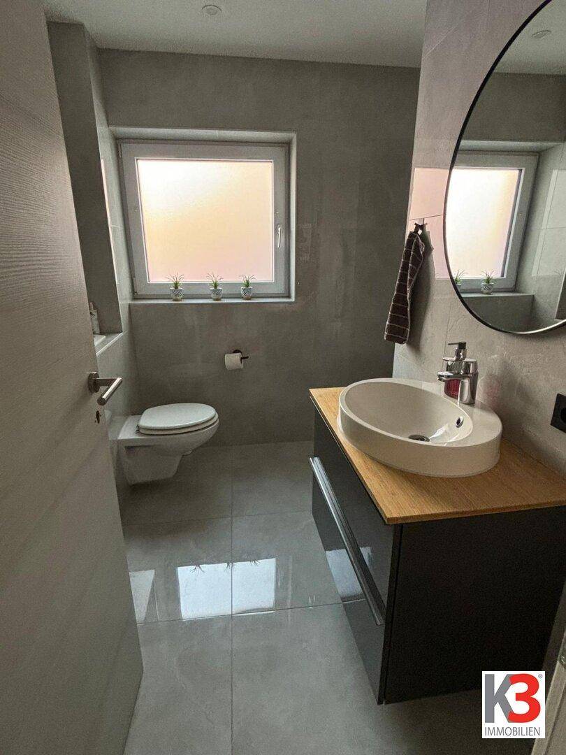 Modernes Gäste-WC mit Waschtisch, Wand-WC und Fenster, in grauen Tönen gehalten.