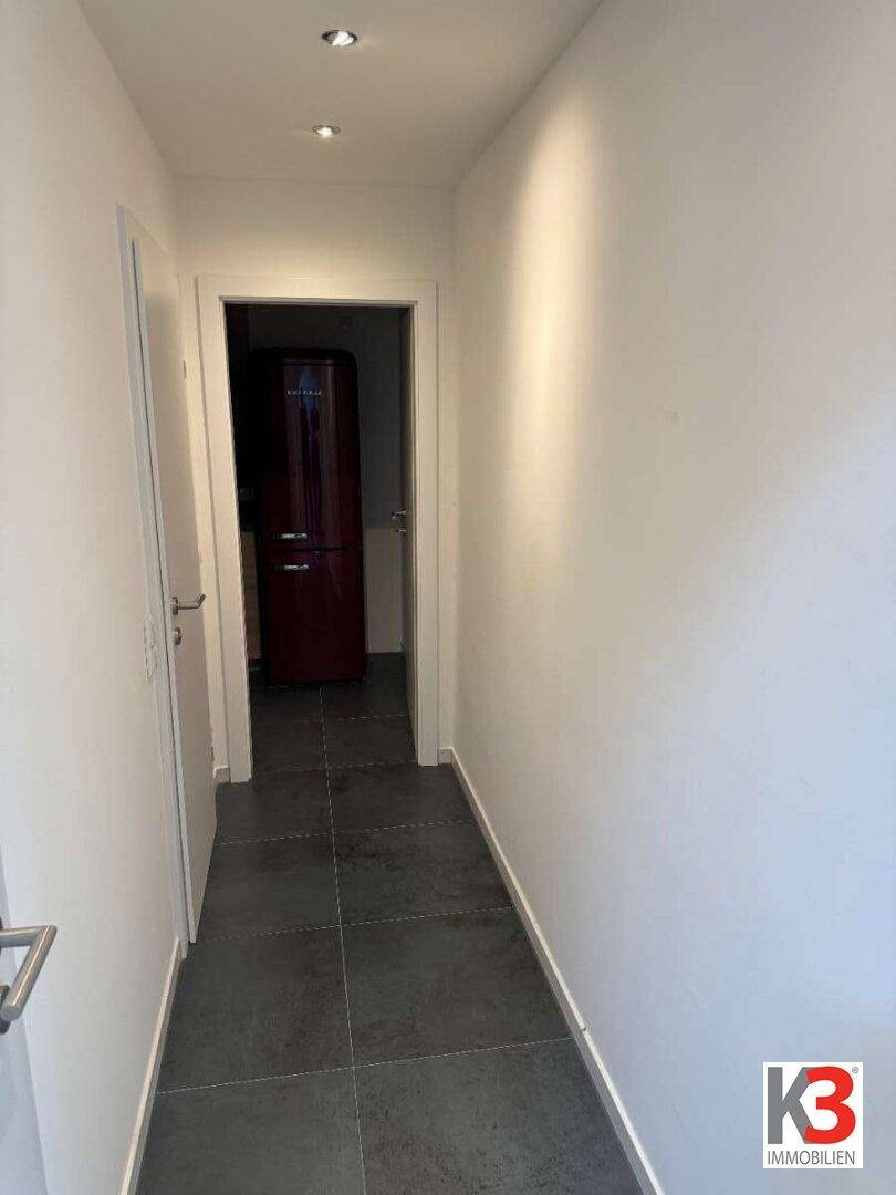 Langer Flur mit dunklen Fliesen und Zugang zu verschiedenen Zimmern, inklusive Kühlschrank.