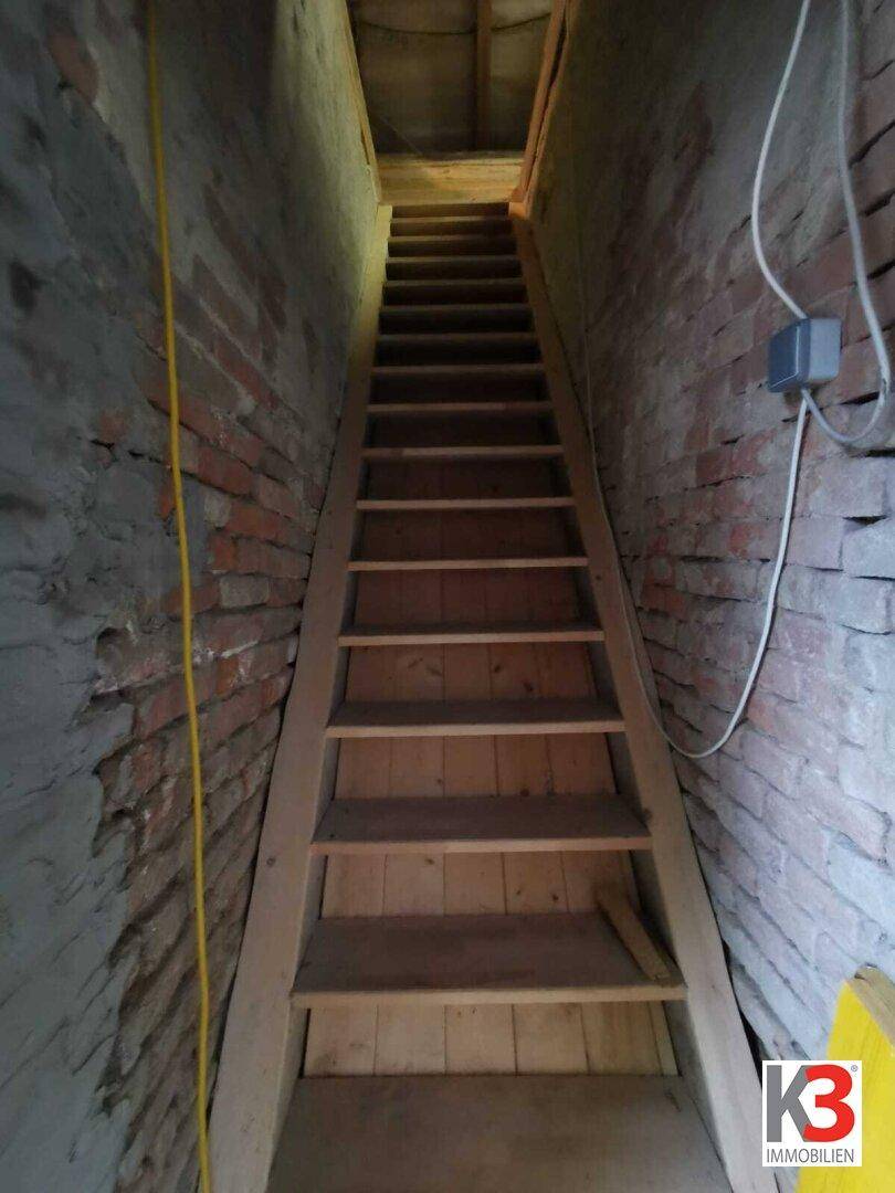 Eine Holztreppe mit unverputzten Ziegelwänden, die in ein Obergeschoss führt.