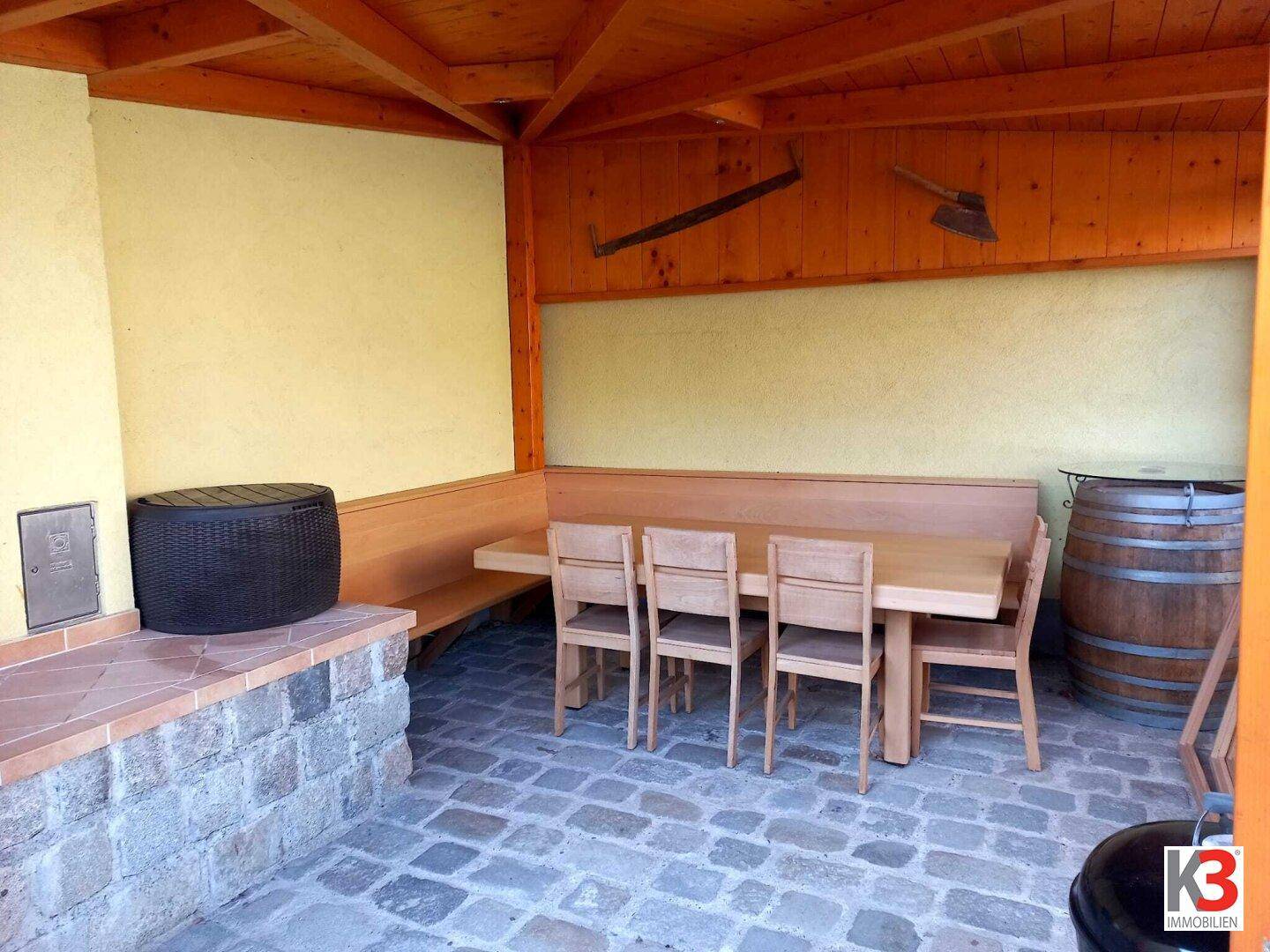 Die überdachte Terrasse mit Holztisch, Bänken und einem gemauerten Grillbereich.