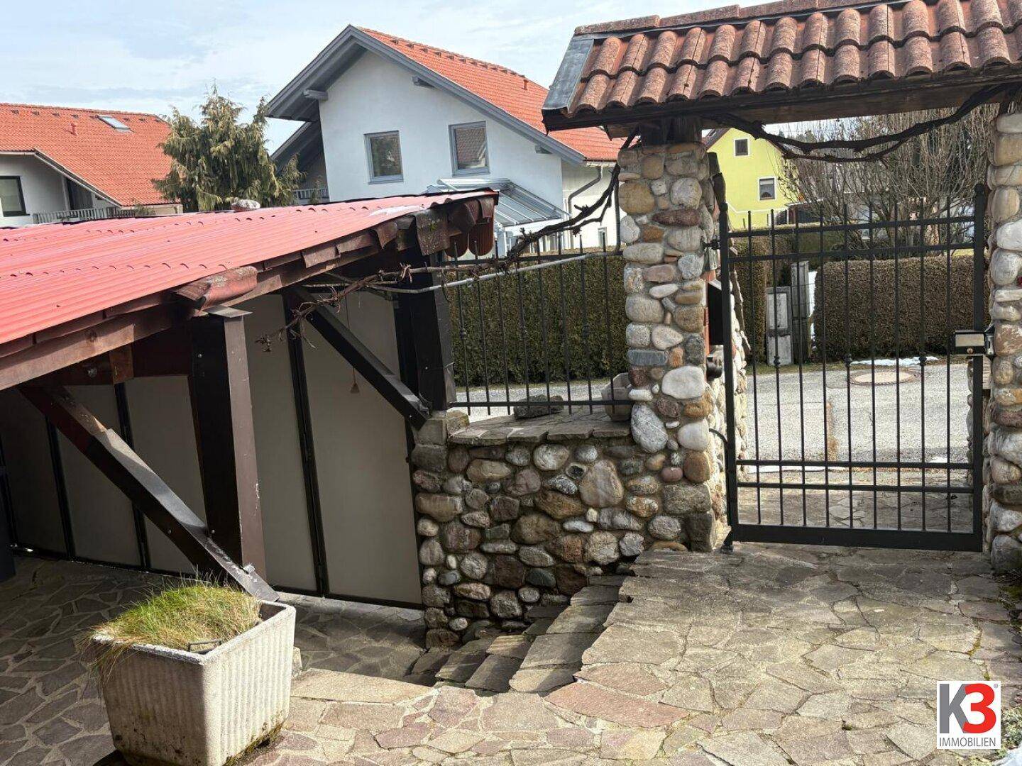 Außenbereich mit Steinfassade, Einfahrtstor und überdachtem Stellplatz für Fahrzeuge.