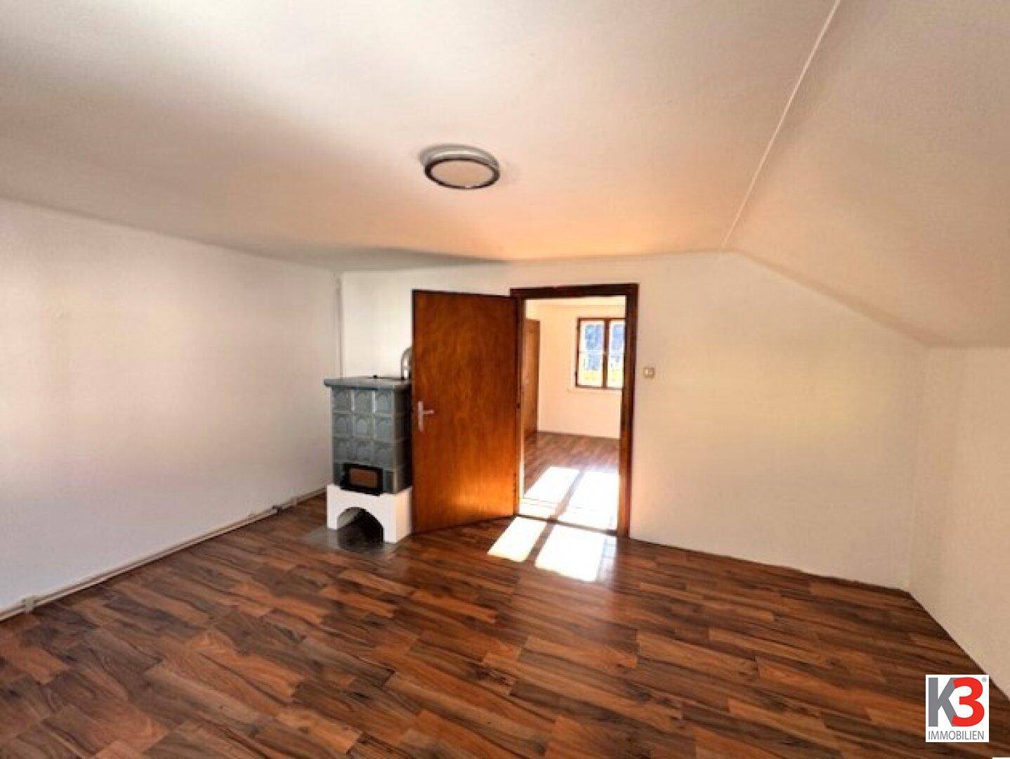 Gemütliches Zimmer mit Holzboden und traditionellem Kachelofen, durch eine offene Tür verbunden.