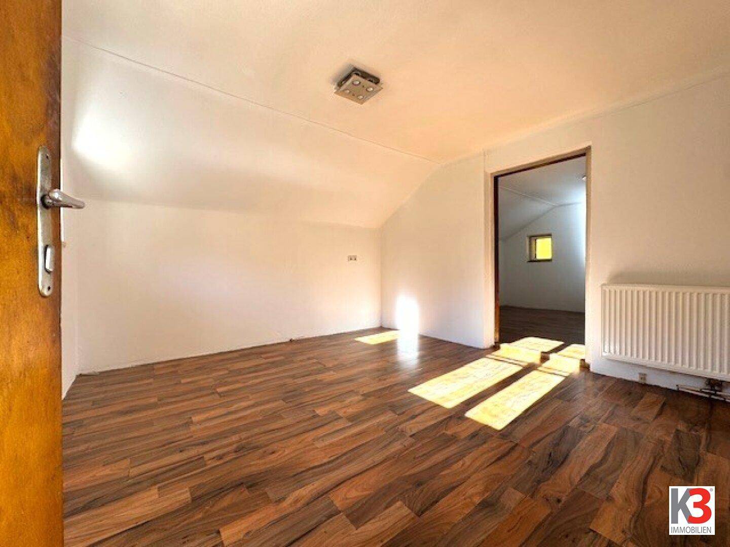 Heller Raum mit Holzboden und Dachschräge, durchflutet von natürlichem Sonnenlicht.