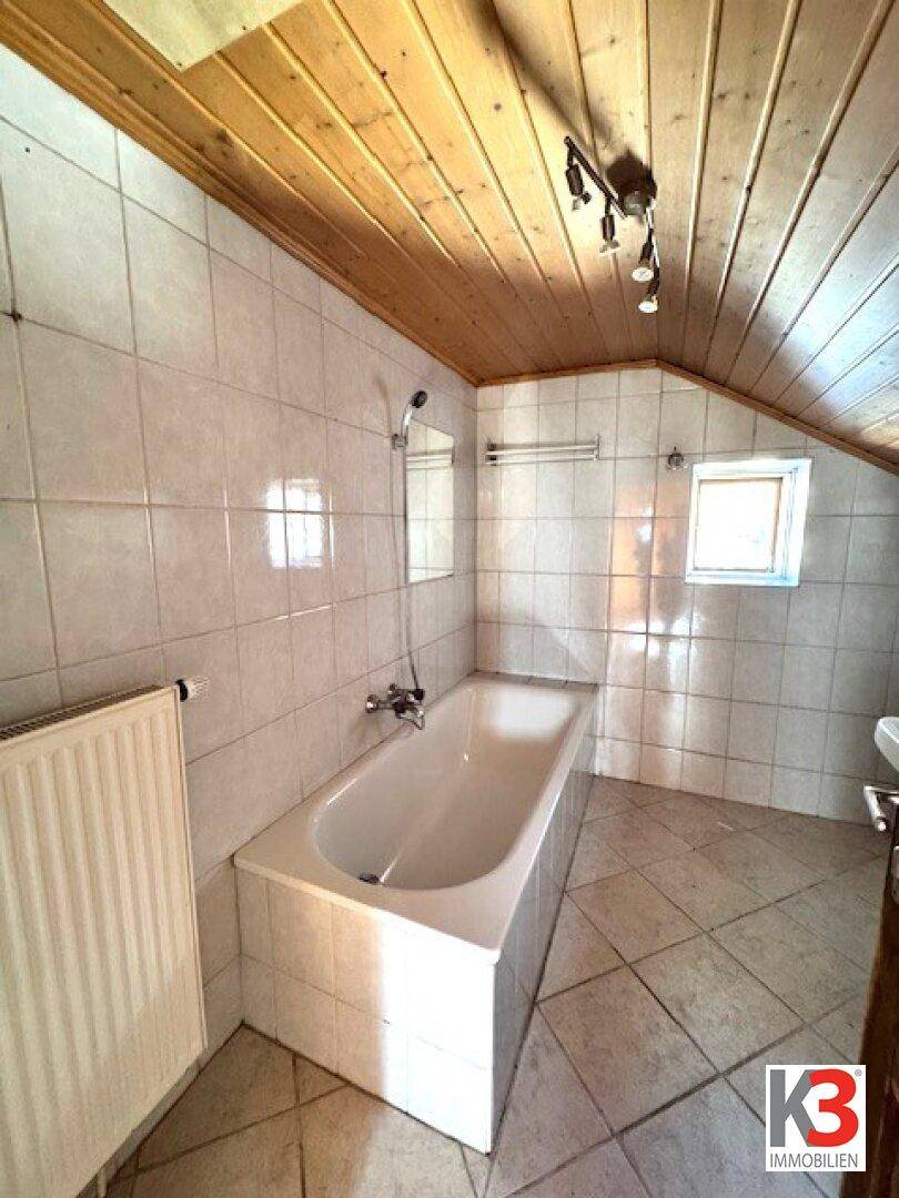 Badezimmer mit Badewanne, Fliesenboden und Holzdecke unter einer Dachschräge.