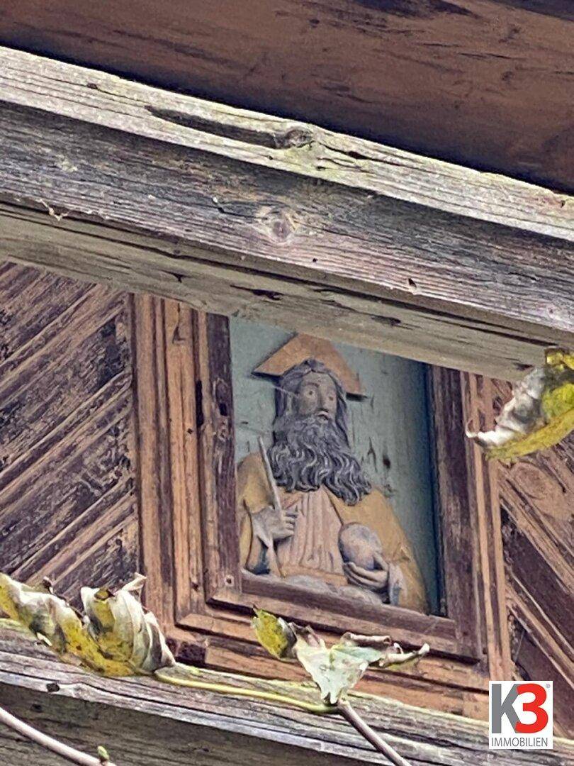 Nahaufnahme einer alten, verwitterten Holzvertäfelung mit einer geschnitzten Figur, umgeben von herbstlichen Blättern.