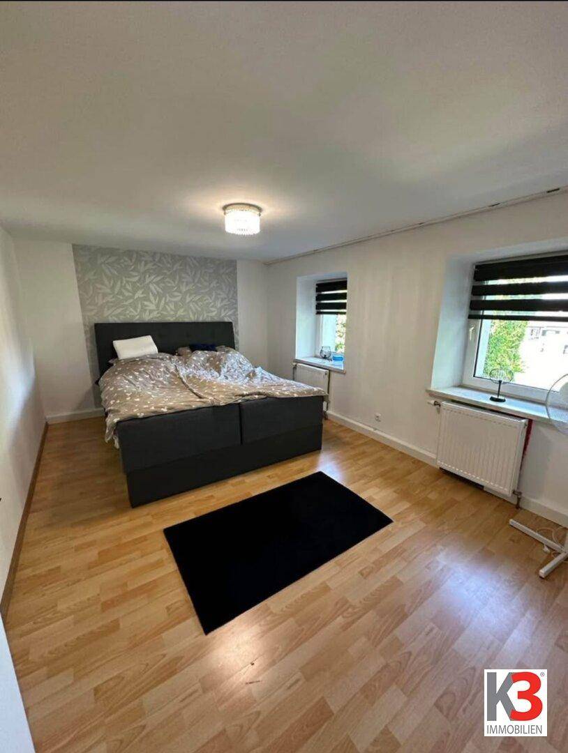 Helles Schlafzimmer mit Doppelbett, Holzparkett und zwei Fenstern mit Jalousien.