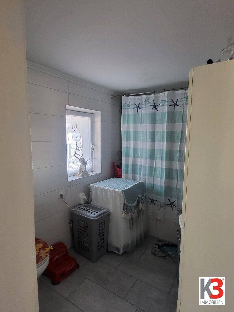 Badezimmer mit Duschvorhang, Fenster und Waschkorb, hell gefliest.