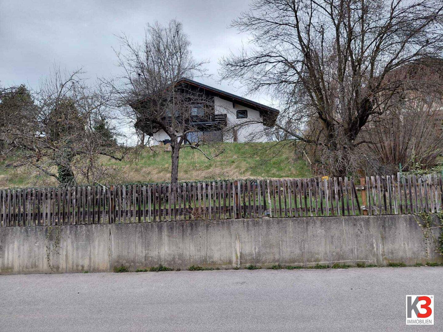 Haus auf einem Hügel, sichtbar hinter einem Holzzaun und einer Betonmauer, mit kahlen Bäumen.