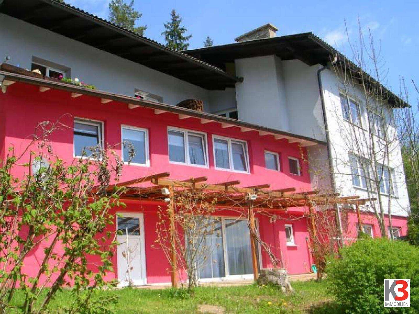 Rote Gebäudefassade mit großen Fenstern und einer Holzpergola im Gartenbereich, umgeben von Grün.
