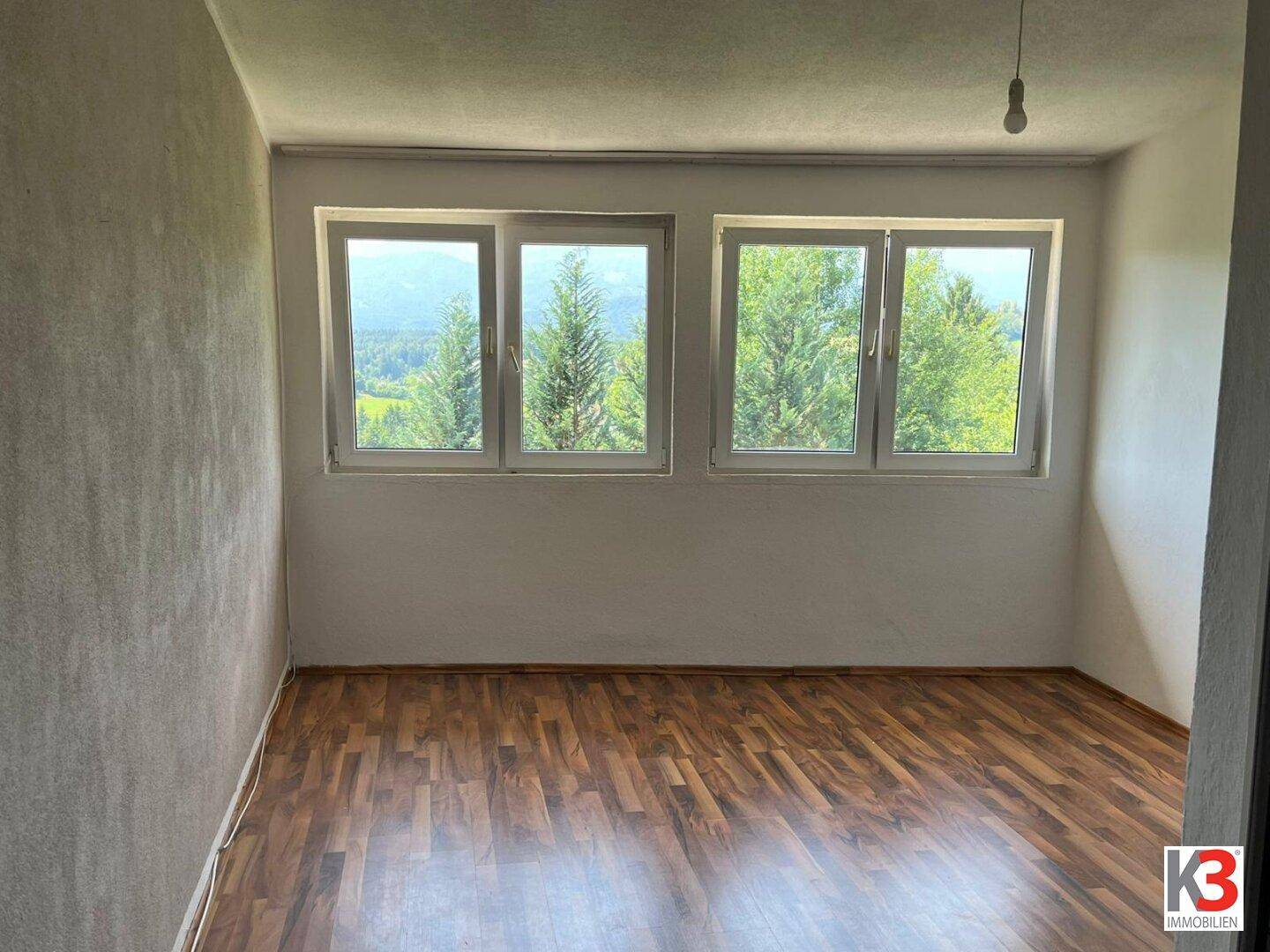 Heller Raum mit Laminatboden und großen Fenstern, die einen weiten Blick ins Grüne bieten.