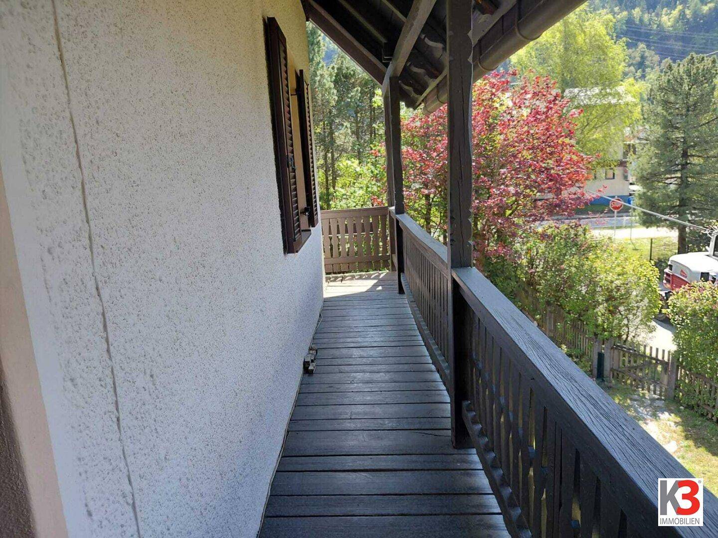 Langer Holzbalkon mit Geländer und Blick auf die umliegende Natur und eine ruhige Straße.