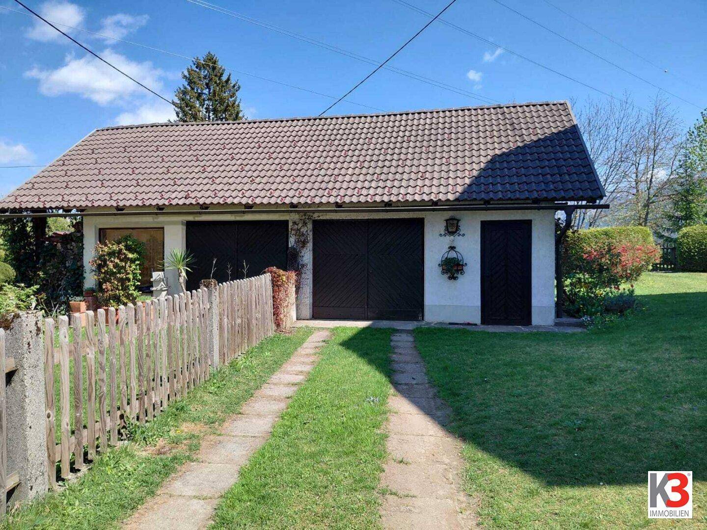 Doppelgarage mit Satteldach und Zufahrt, umgeben von Rasen und einem Holzzaun.