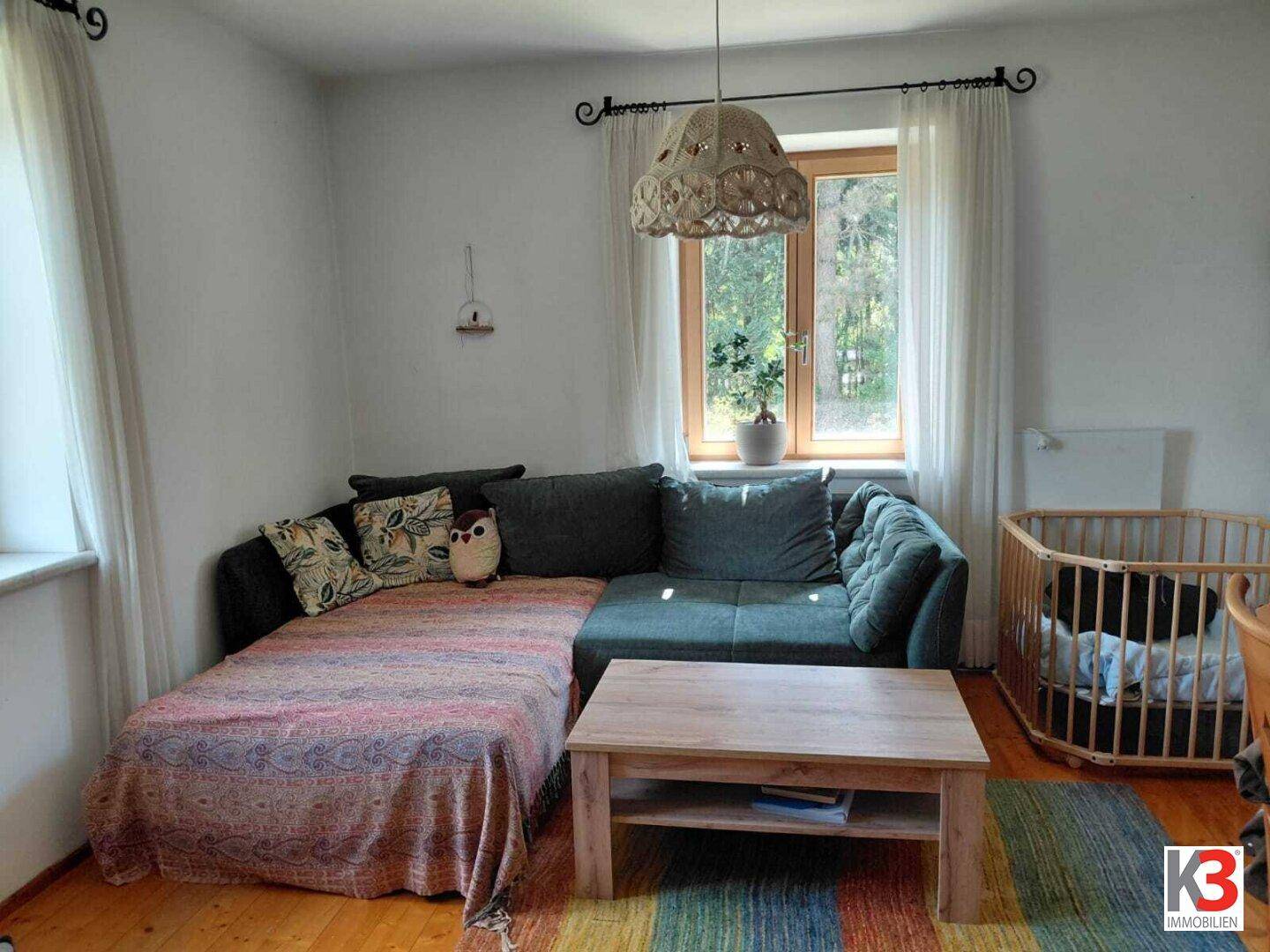 Helles Wohnzimmer mit Ecksofa, Holztisch und Kinderbett, ausgestattet mit bunten Teppichen.