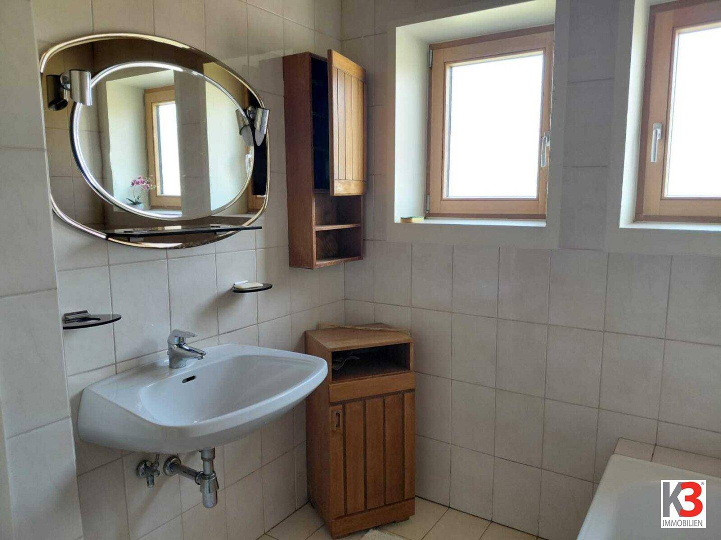 Badezimmer mit Waschbecken, Spiegelschrank und hellen Fliesen, gut beleuchtet durch Fenster.