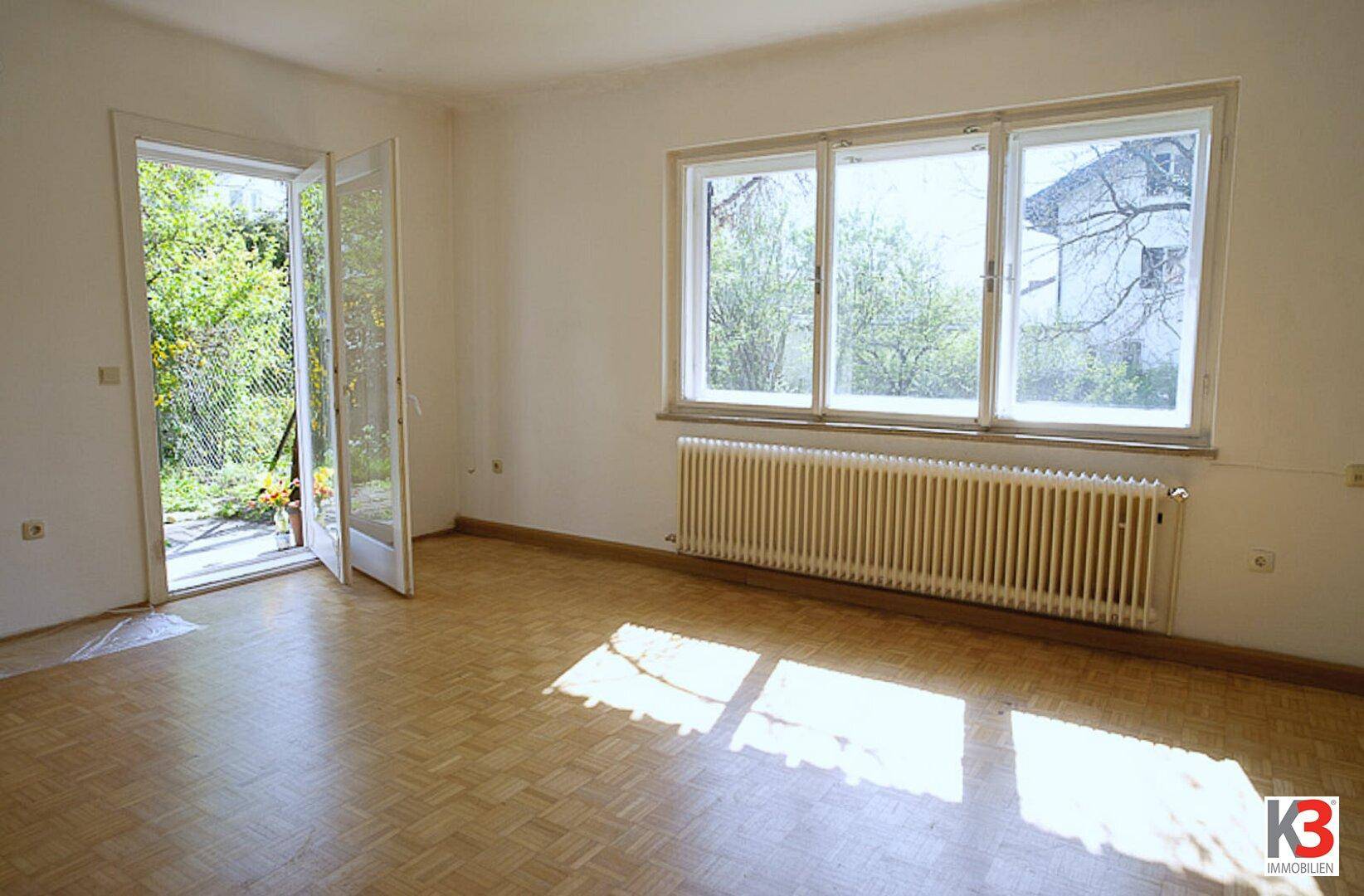 Helles, leeres Zimmer mit Parkettboden, großem Fenster und direktem Zugang zum Gartenbereich.