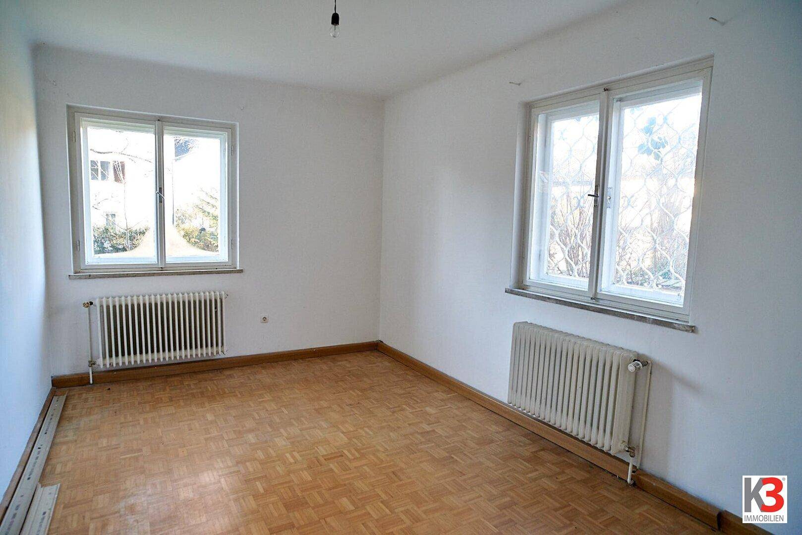 Helles Schlafzimmer mit Parkettboden, zwei Fenstern und Heizkörpern, ideal für individuelle Einrichtung und Komfort.