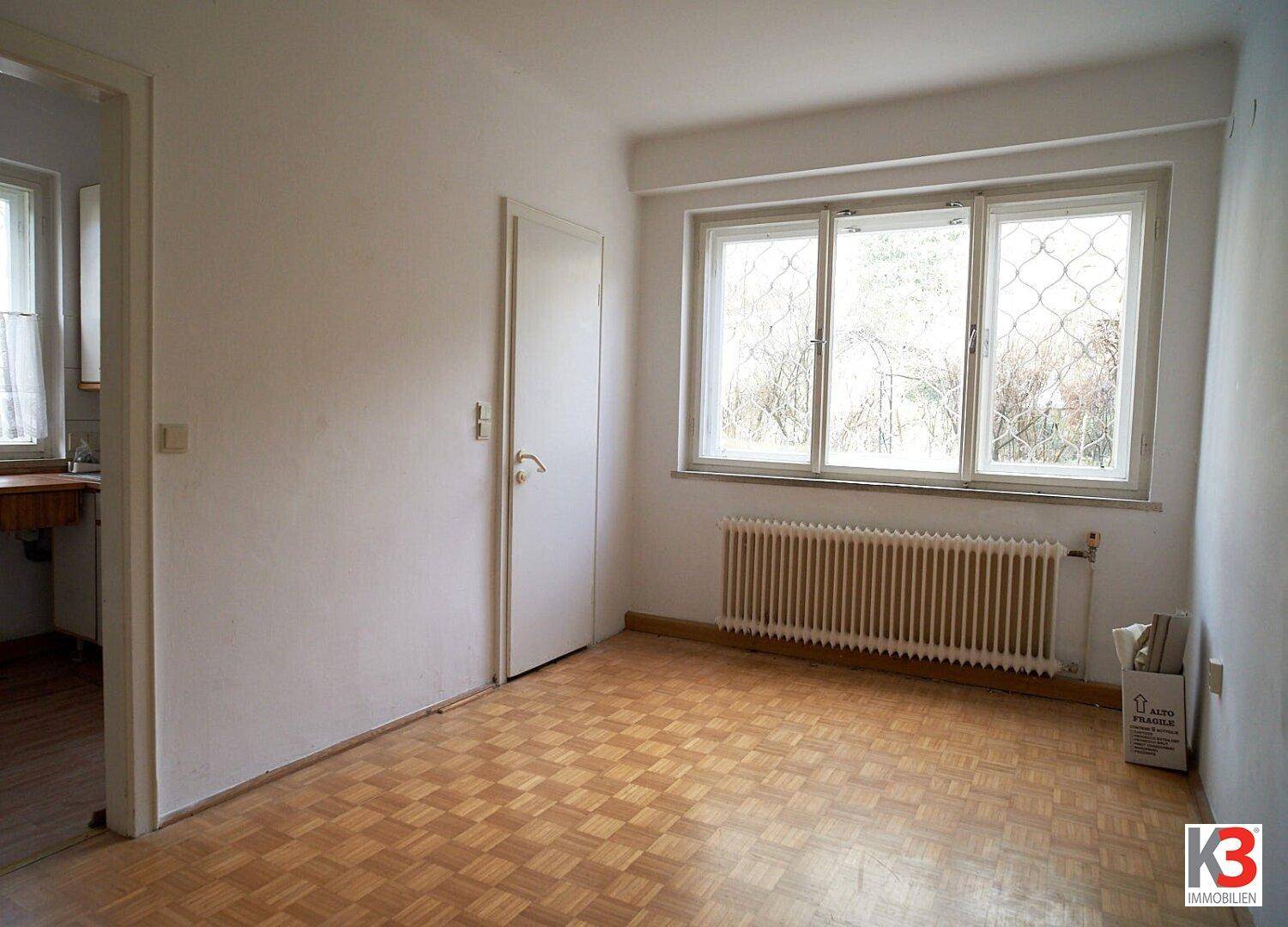 Leeres Zimmer mit Parkettboden, Fenster und zwei Türen, bereit zur individuellen Gestaltung und Nutzung.