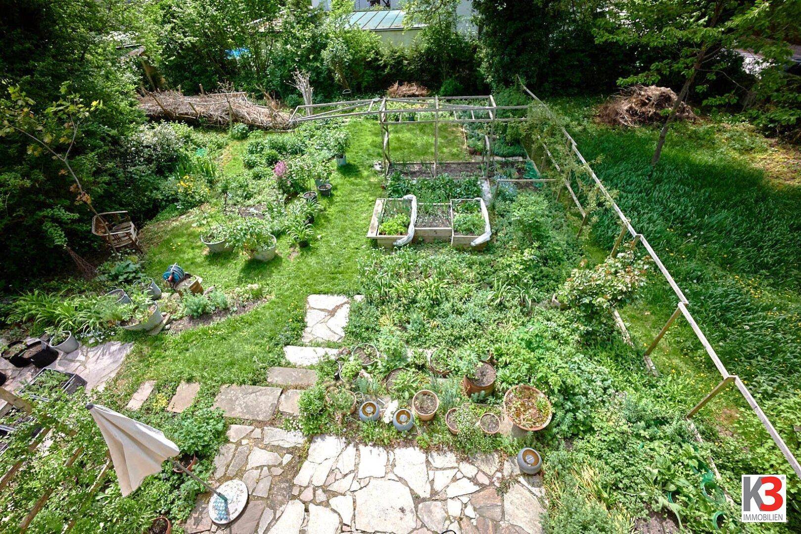 Weitläufiger, üppiger Garten mit Steinterrasse, Hochbeeten und vielfältiger Bepflanzung, von oben betrachtet.