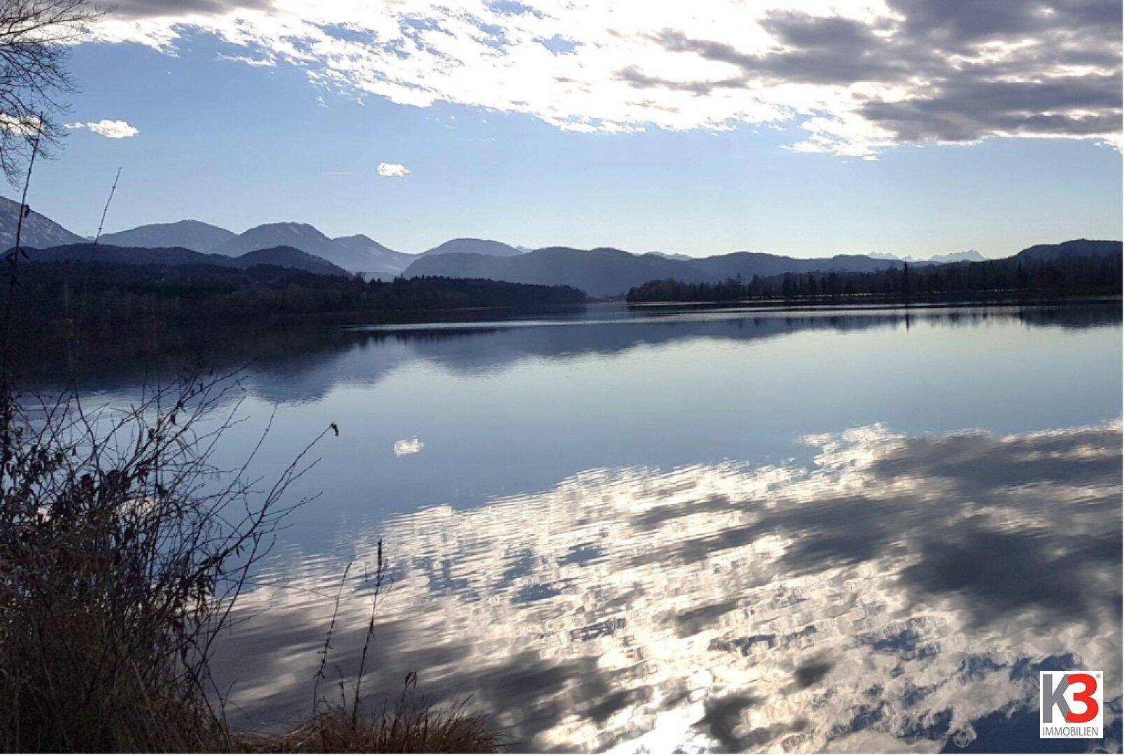 Malerischer Blick auf einen ruhigen See mit majestätischen Bergen im Hintergrund unter einem bewölkten Himmel.