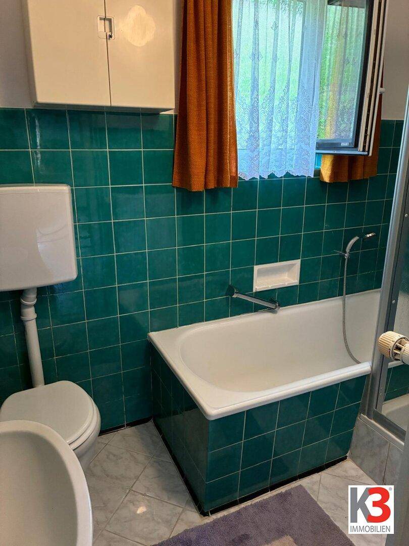 Badezimmer mit grünen Fliesen, Badewanne, Toilette und Fenster mit orangefarbenen Vorhängen.