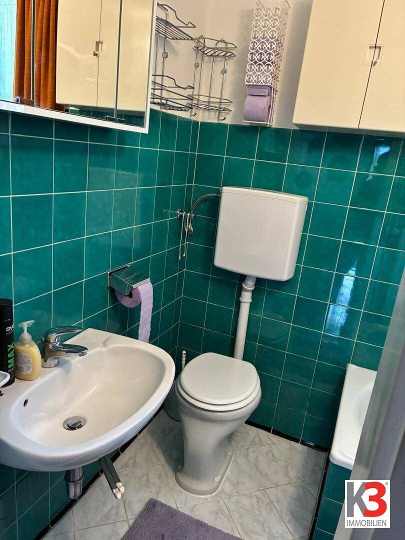 Badezimmer mit grünen Fliesen, Waschbecken, Toilette und Spiegelschrank über dem Waschbecken.