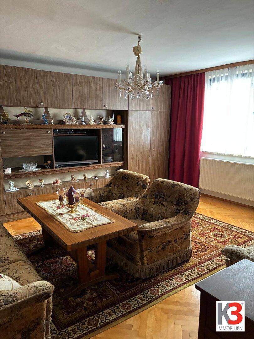 Traditionelles Wohnzimmer mit gemusterten Sesseln, Couchtisch, großer Schrankwand und Parkettboden.