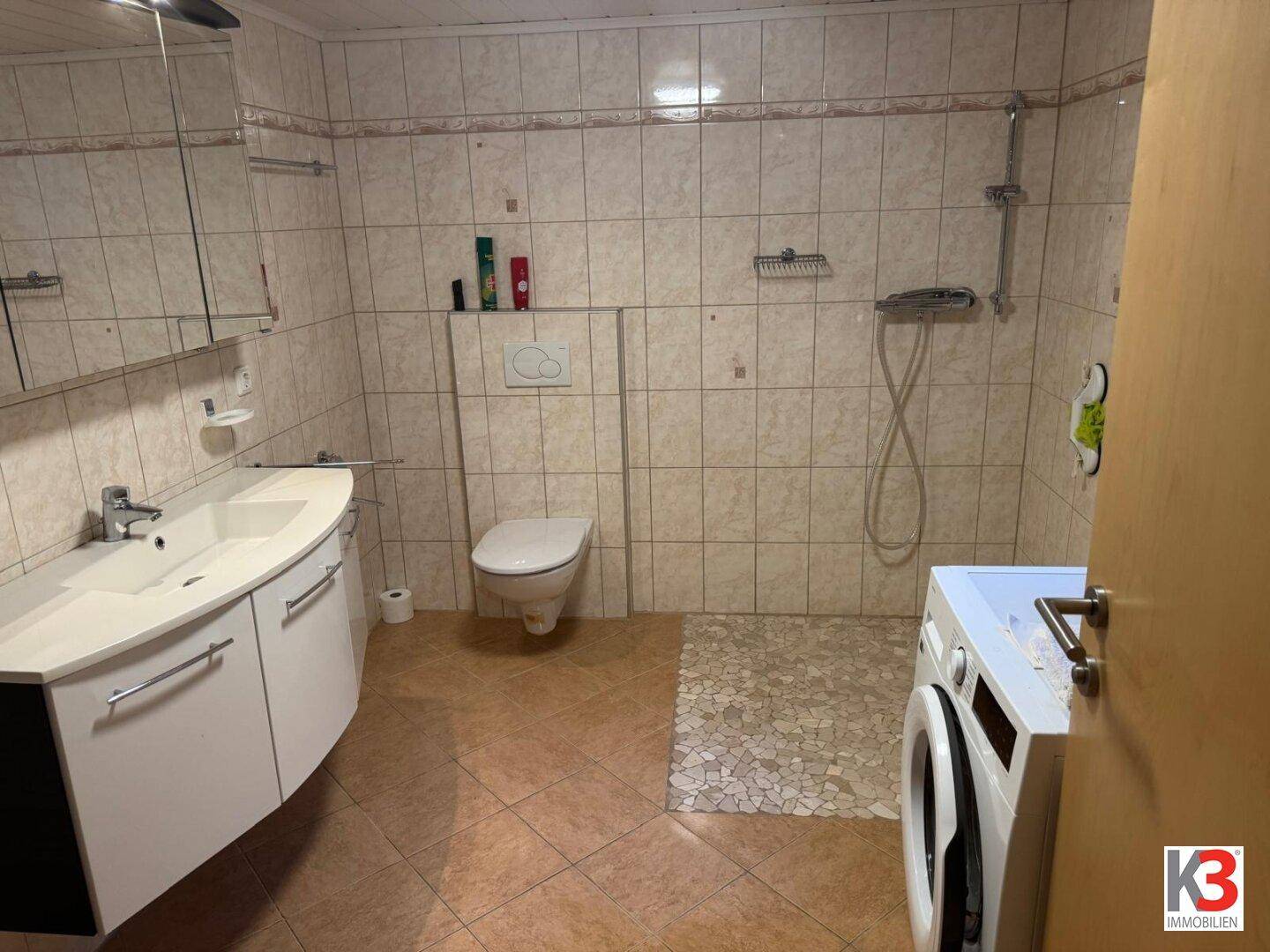 Funktionales Badezimmer mit Fliesen, Waschbecken, WC und integrierter Dusche mit Waschmaschine.