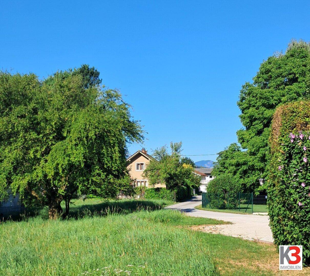 Ländliche Umgebung mit einem Haus im Hintergrund, umgeben von Bäumen und einer weiten Grasfläche unter blauem Himmel.