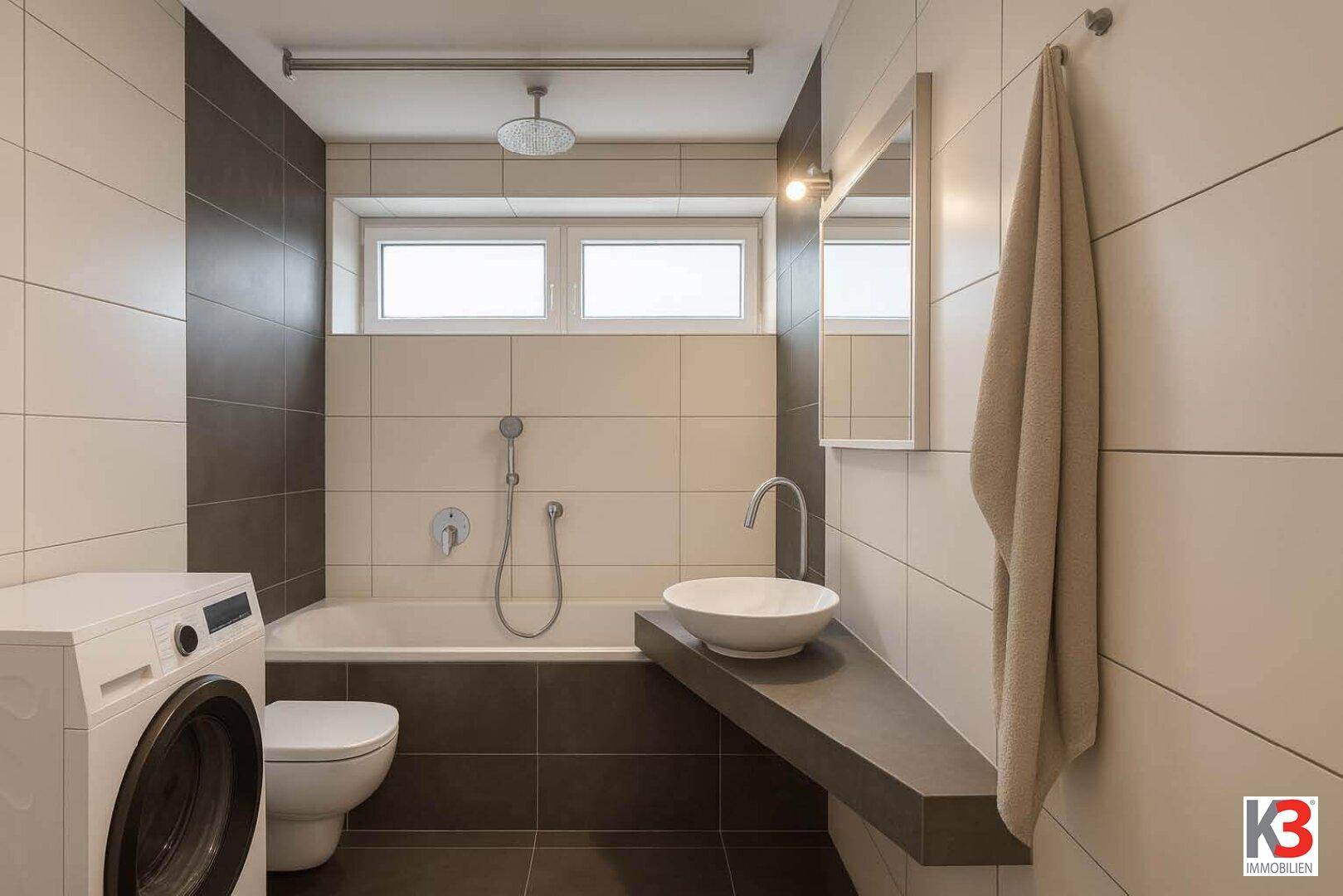 Modernes Badezimmer mit Badewanne, Waschmaschine, stilvollem Waschbecken und Fliesen in zwei Farbtönen.