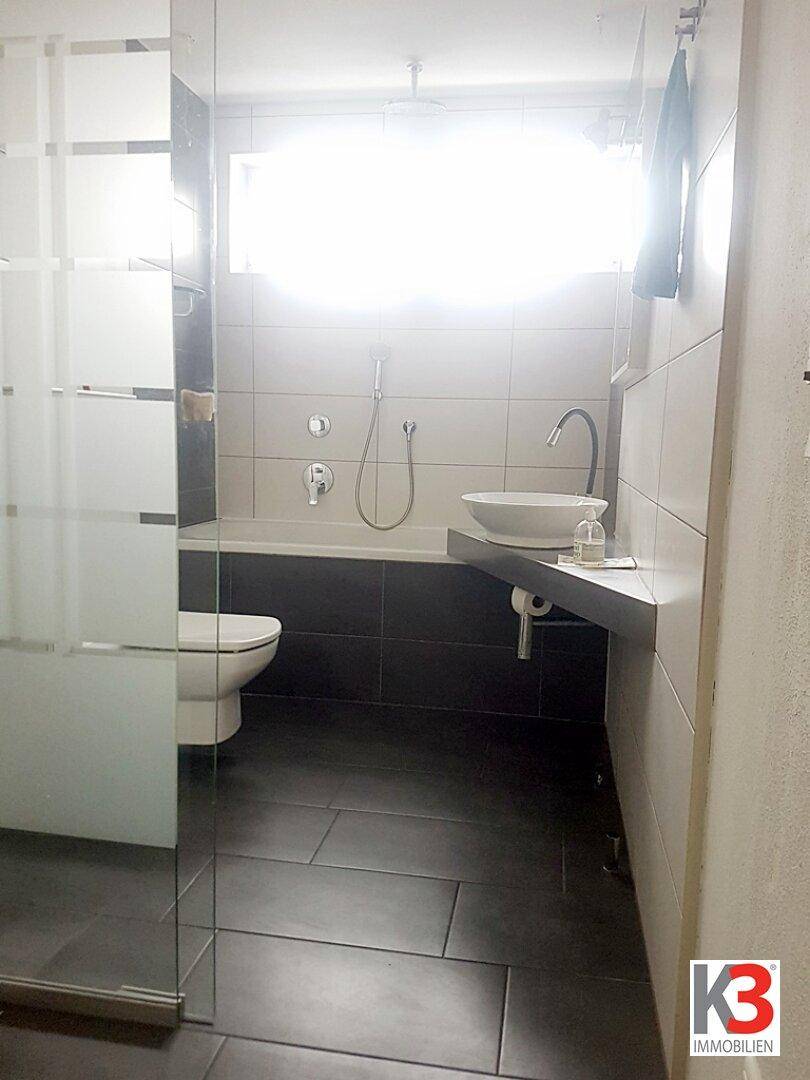 Helles Badezimmer mit Badewanne, WC, modernem Waschbecken und einer Duschabtrennung aus Glas.