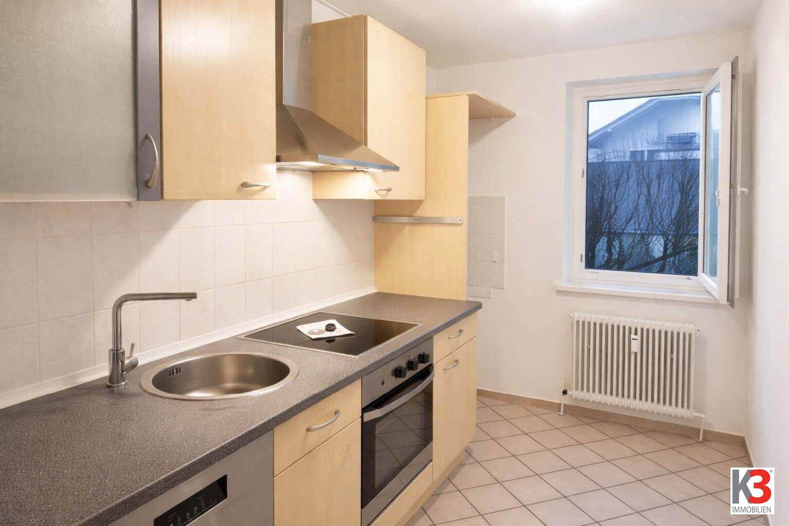 Funktionale Küche mit hellen Holzschränken, dunkler Arbeitsplatte und einem Fenster, das Tageslicht spendet.
