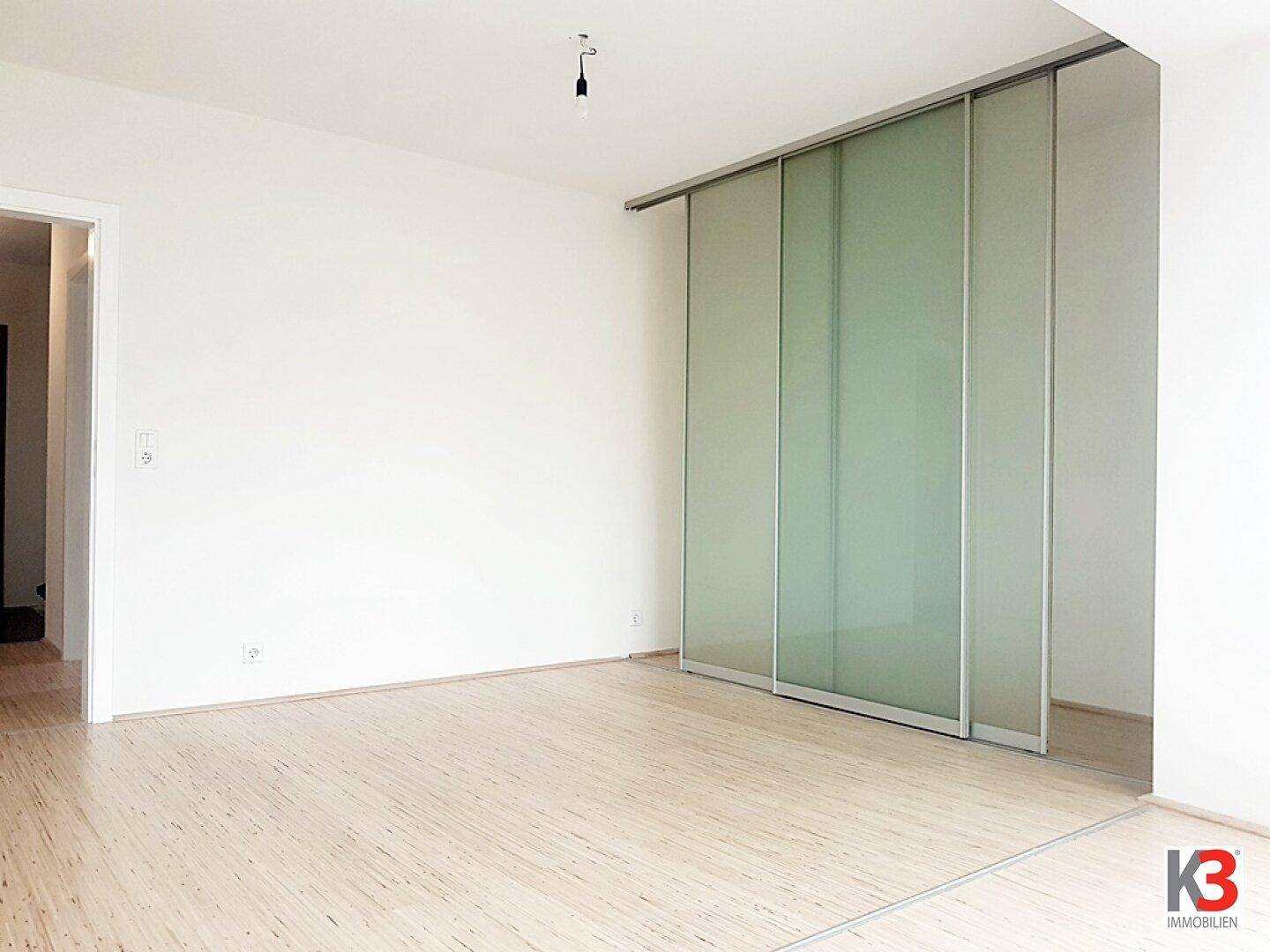 Leerer Raum mit hellen Wänden, Holzboden und einer modernen Glasschiebetür, die den Bereich abtrennt.