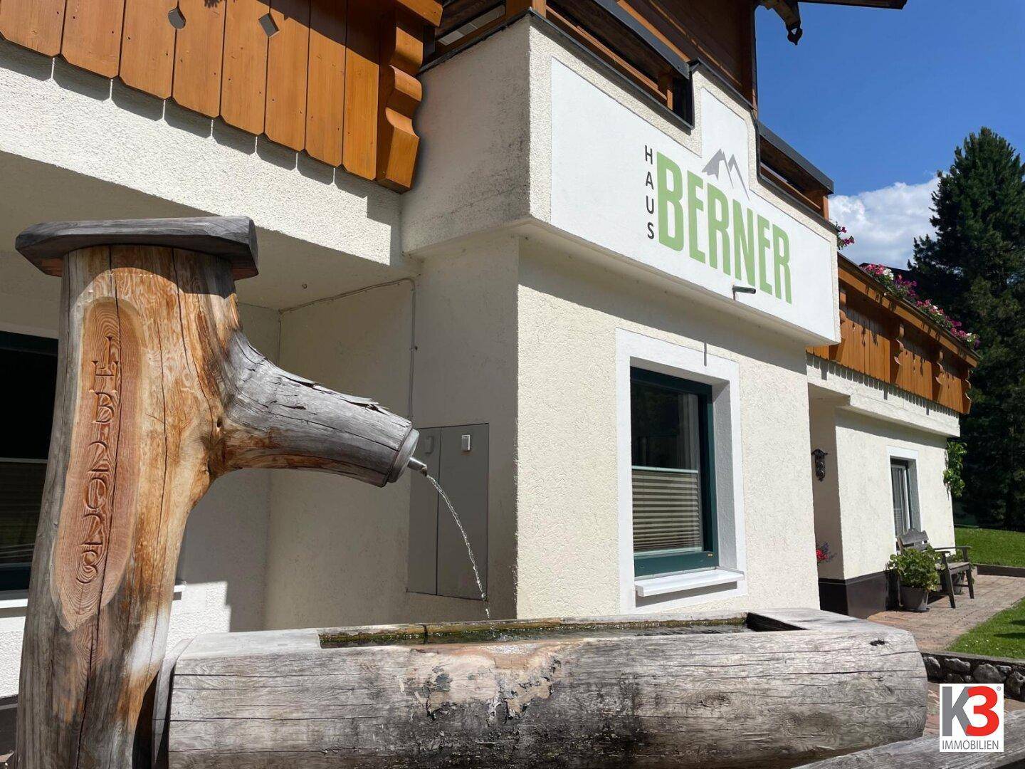 Eingangsbereich des Gebäudes mit einem rustikalen Holzbrunnen und Bergblick.