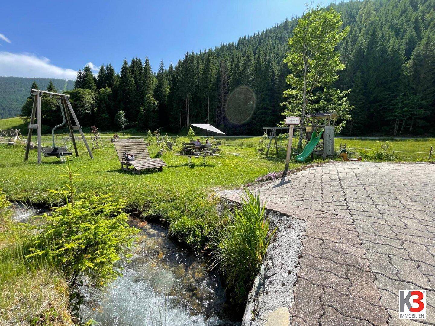 Spielbereich im Freien mit Bachlauf, Schaukeln und Sitzgelegenheiten in der Natur.