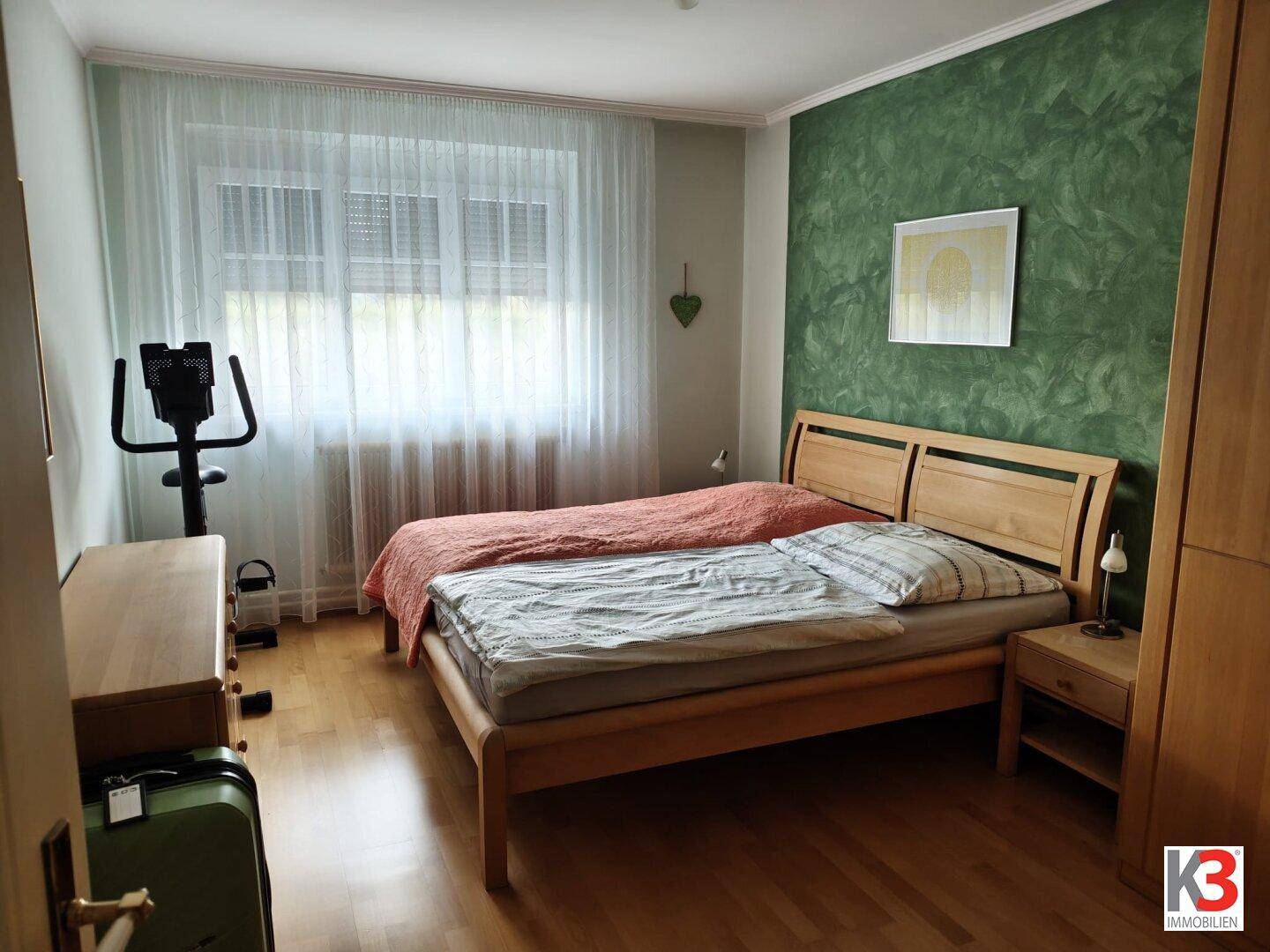Schlafzimmer mit Holzbett, grüner Akzentwand und großem Fenster für viel Tageslicht.