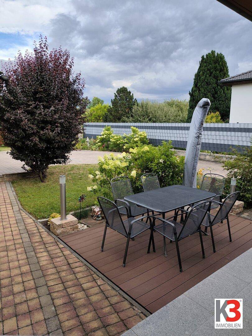 Gemütliche Holzterrasse mit Esstisch und Gartenmöbeln, umgeben von einem gepflegten Garten unter bewölktem Himmel.