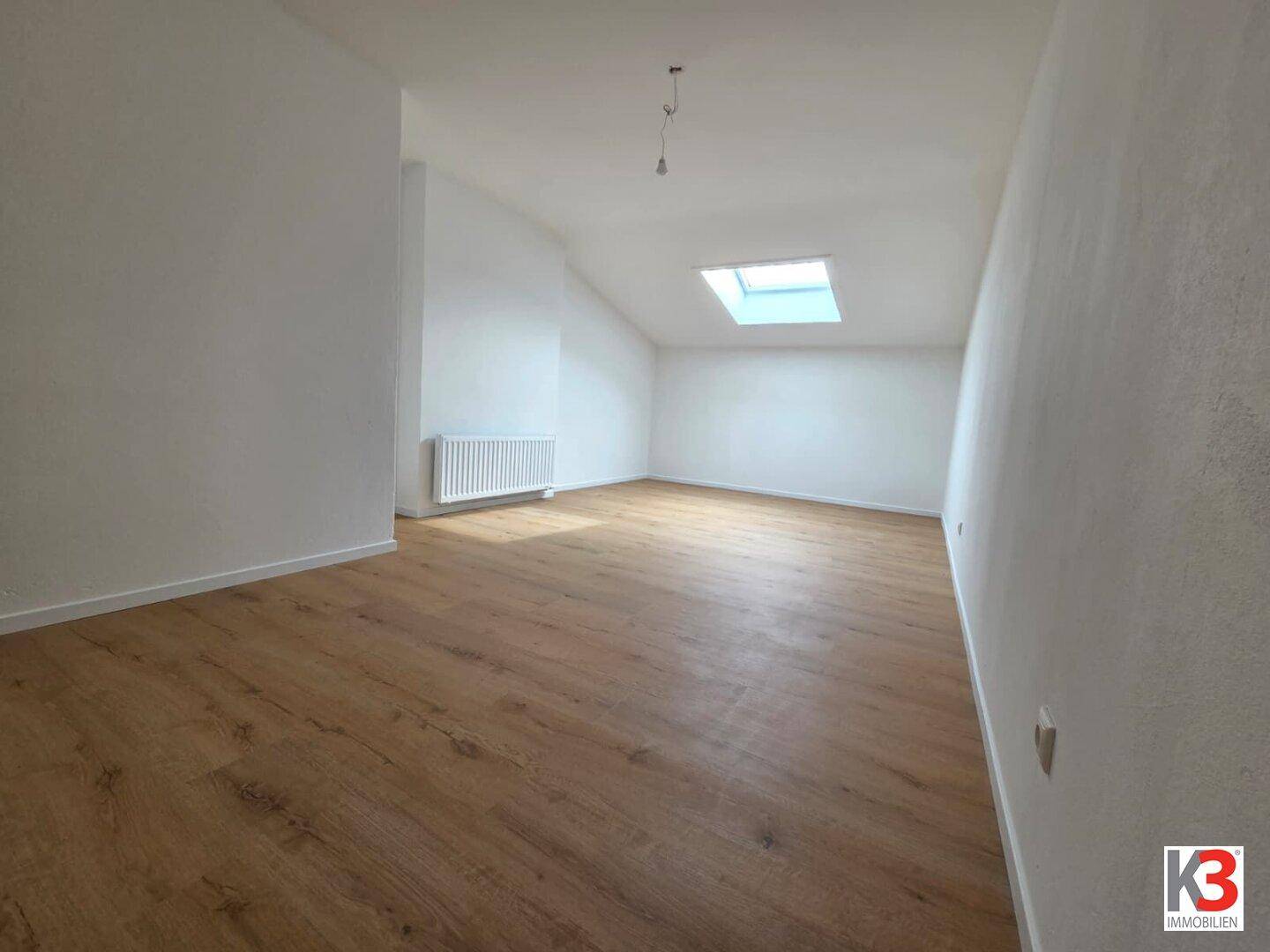 Heller, leerer Raum mit Laminatboden, weißen Wänden und einem Dachfenster.