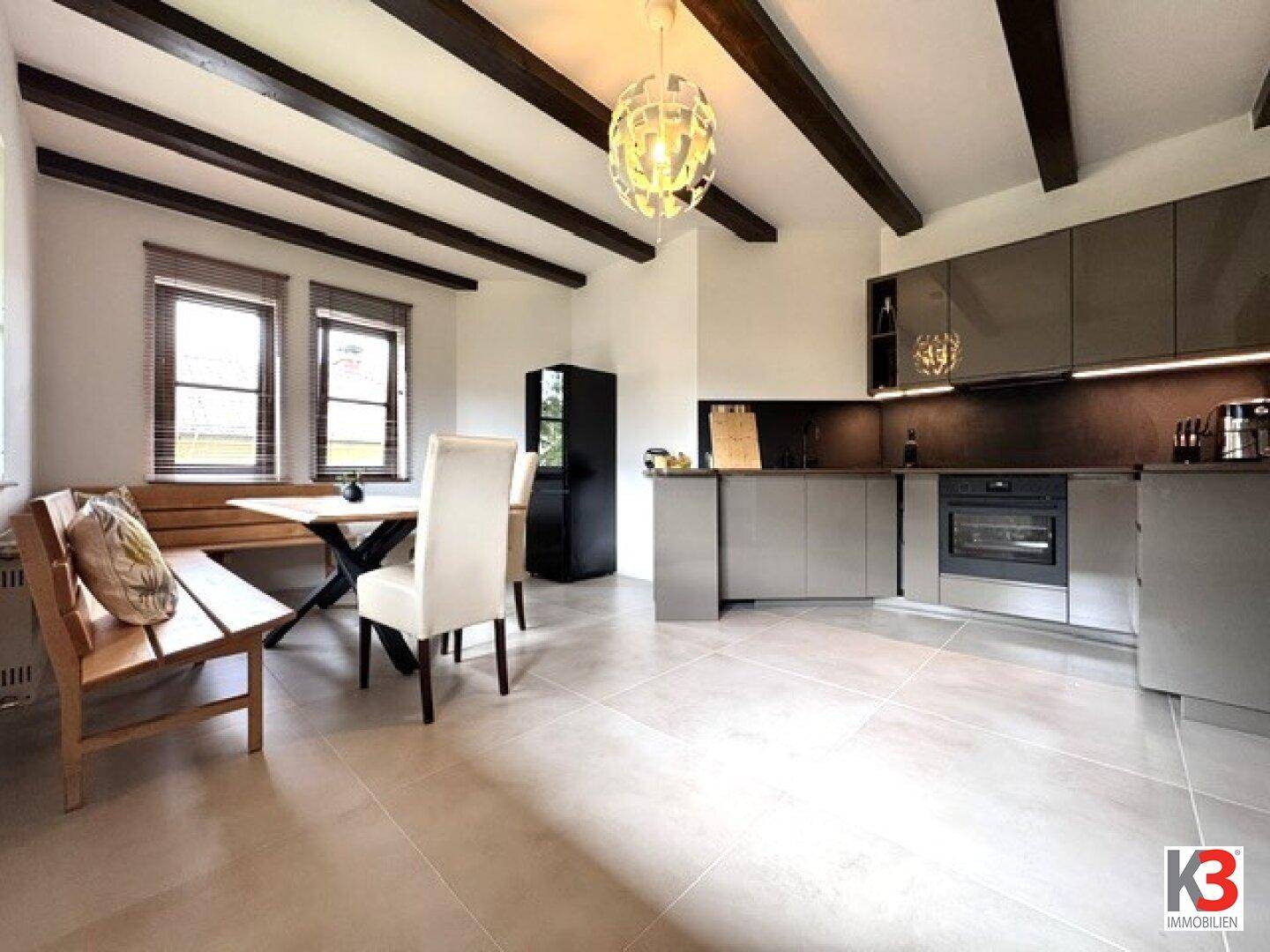 Moderne Küche mit Essbereich, grauen Schränken, sichtbaren Holzbalken und Fliesenboden.